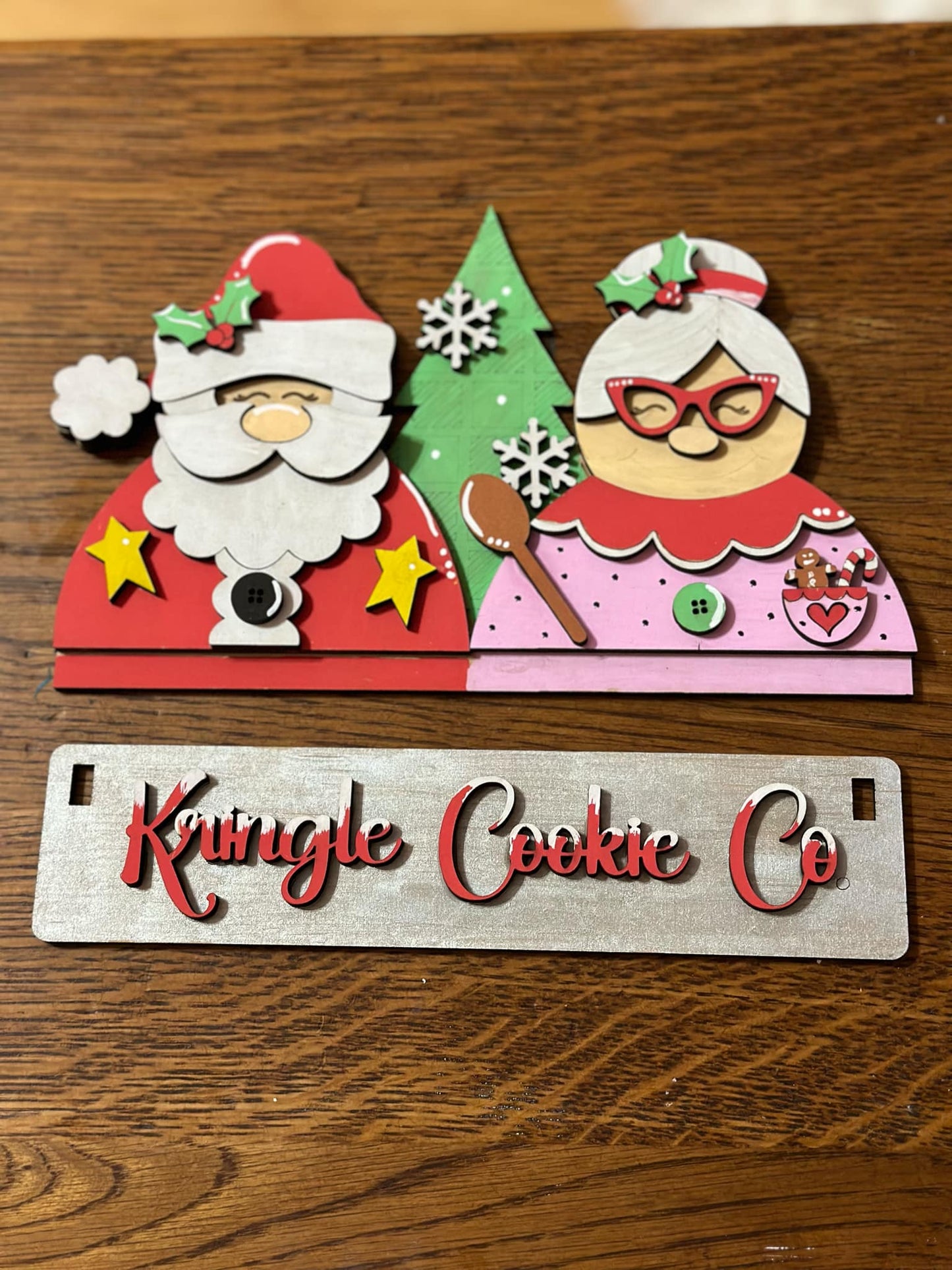Laser-cut DIY MDF Christmas craft kit featuring Santa Claus, Mrs. Claus, green Christmas tree, and layered “Kringle Cookie Co.” sign with festive red, pink, and white holiday accents.