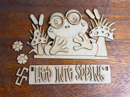 Unfinished Hop Into Spring Frog DIY craft kit with laser-cut wood pieces including frog, mushrooms, flowers, and interchangeable spring sign ready to paint and assemble.