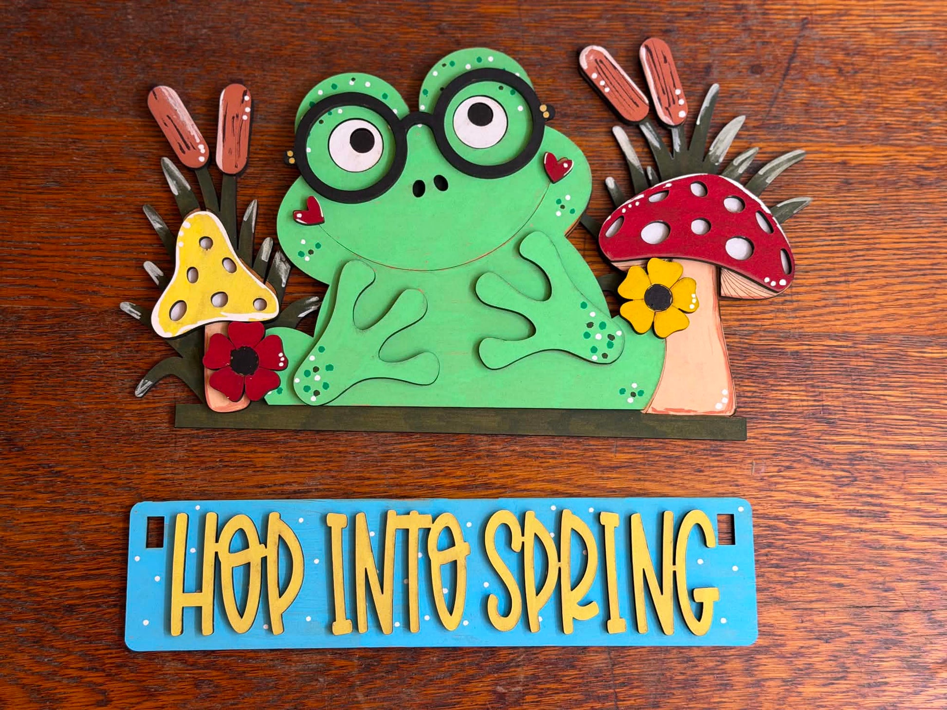 Hand-painted Hop Into Spring Frog DIY craft kit featuring a green frog with glasses, mushrooms, flowers, and interchangeable spring sign for seasonal home decor.