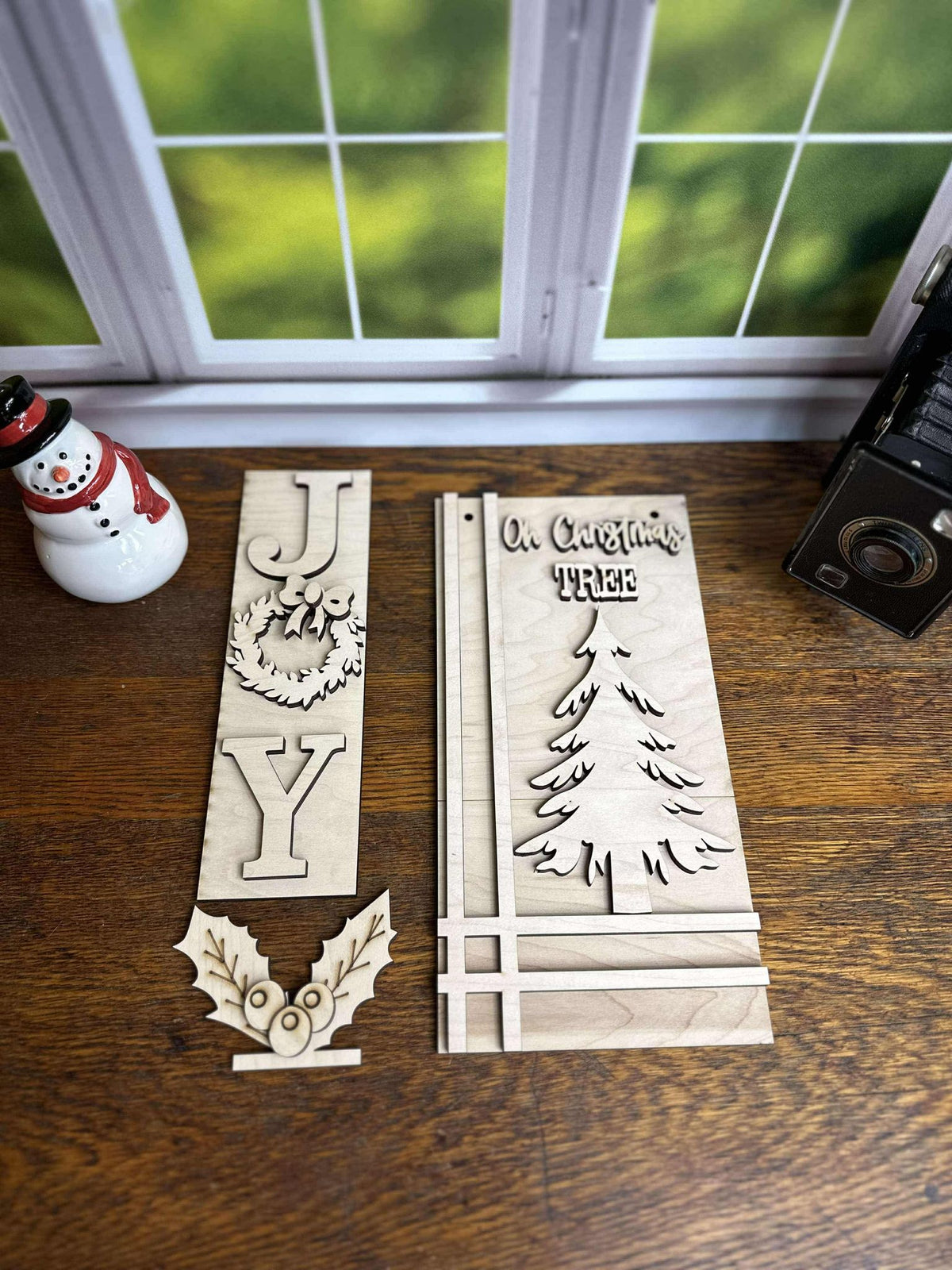 Three-piece JOY Christmas post insert kit arranged on a wooden table, showing the unfinished wreath, Christmas tree sign and holly accent.