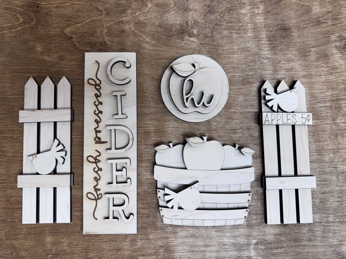 Close-up of unfinished laser-cut apple orchard décor pieces including cider sign, apple basket and wooden fence accents