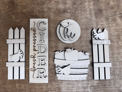 Close-up of unfinished laser-cut apple orchard décor pieces including cider sign, apple basket and wooden fence accents